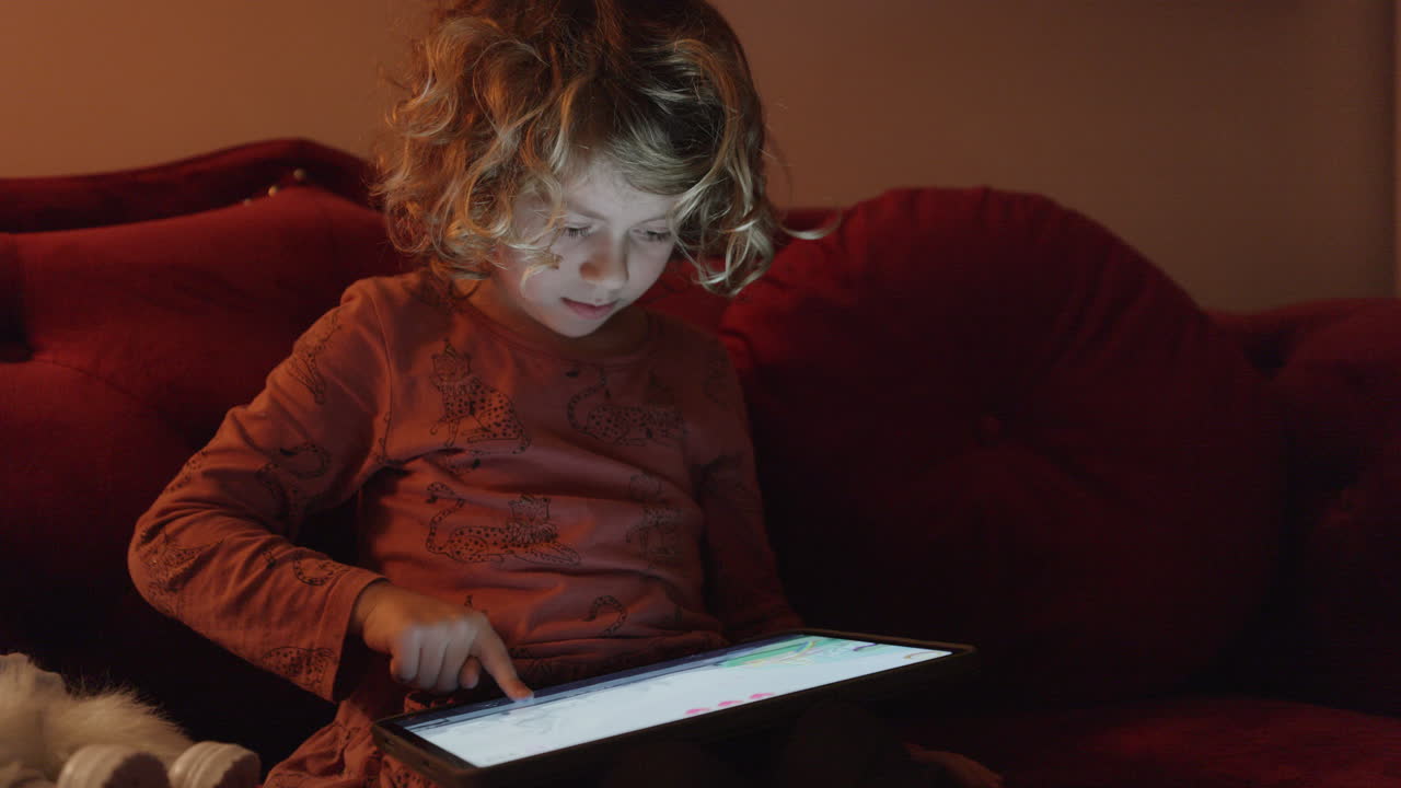 A small girl plays or watches a tablet computer with the light from the screen in a dark room