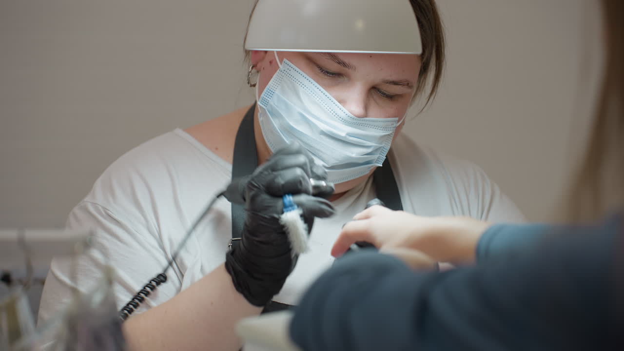 Closeup view of nail technician wearing surgical mask and black gloves using electric nail filer and cleaning brush on client nails under white lamp during detailed manicure session in salon