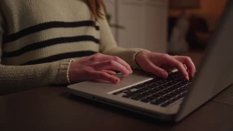 White-female-hands-and-fingers-typing-on-a-silver-laptop-with-a-black-keyboard-on-a-dark-wood-table-in-low-light-at-night-working-from-home