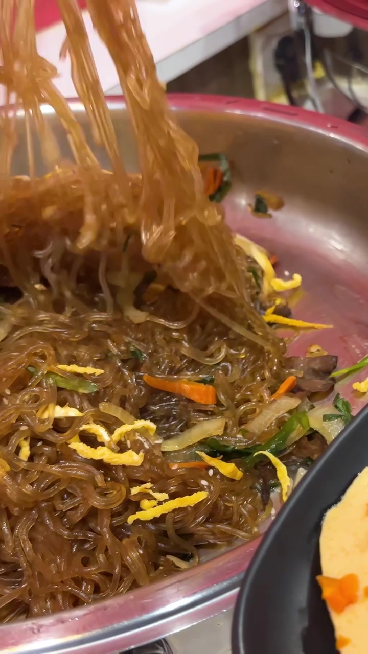 Penjepit mengangkat dan mengaduk mi Japchae berwarna-warni di dapur yang ramai, menonjolkan bahan-bahan yang cerah dan gerakan dinamis.
