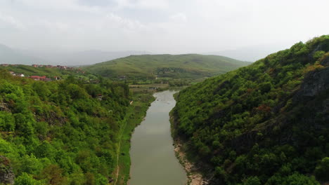 Aerial-push-through-canyon-in-Kosovo,-along-the-Drin-river-as-people-kayak-past-the-town-below