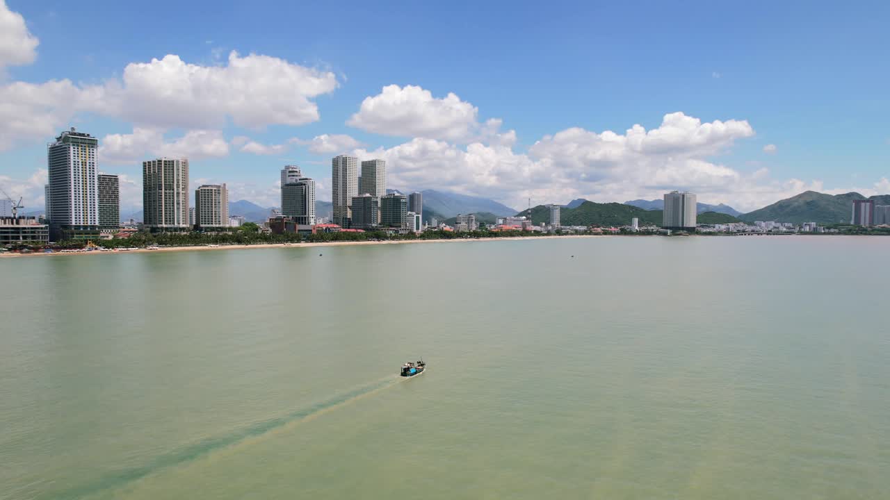 drone aéreo que rodea un barco de pesca que navega en el turbio mar verde turquesa de nha trang vietnam con un gran horizonte residencial y montañas en el horizonte durante un día soleado