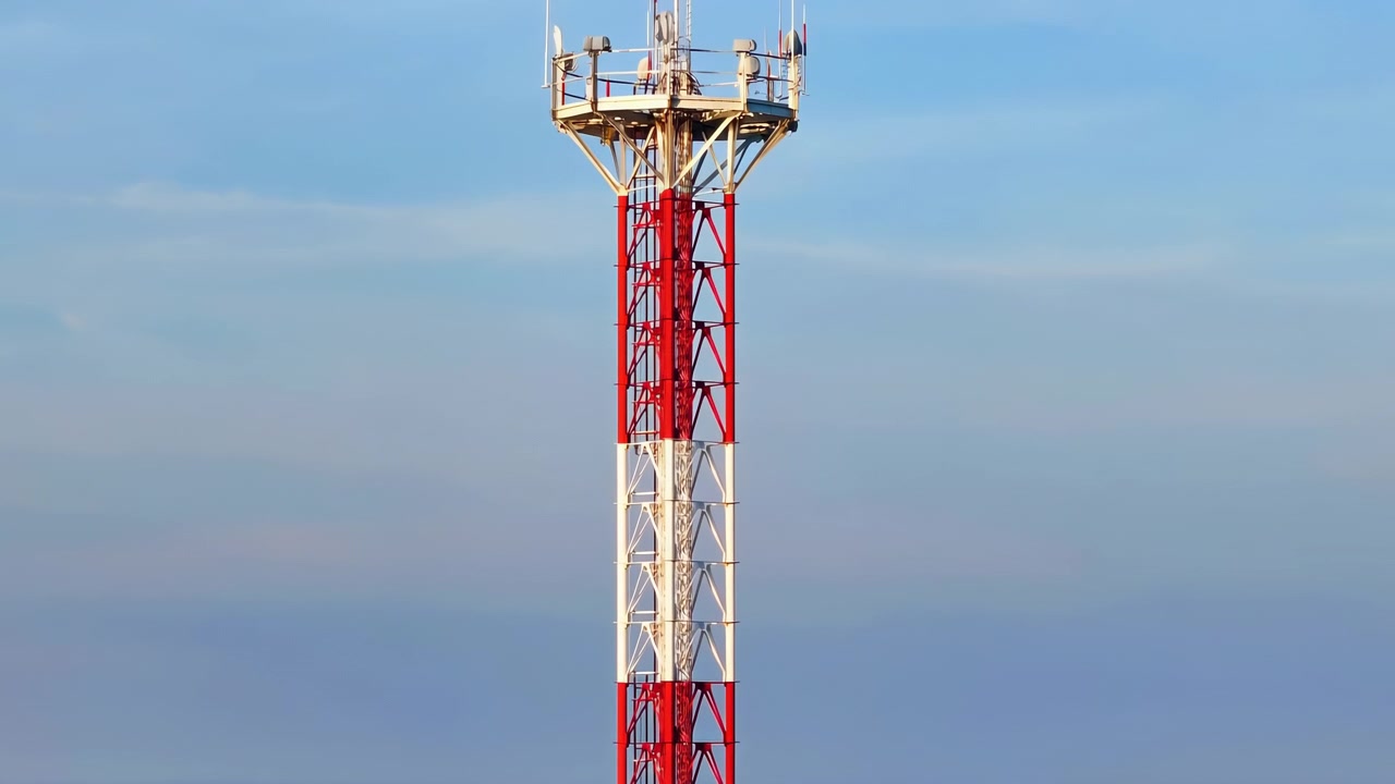 Un alto traliccio di comunicazione a reticolo rosso e bianco si erge centrato contro un cielo azzurro pastello tenue e un orizzonte basso e distante, mostrando la struttura in acciaio, l'infrastruttura di trasmissione, una simmetria pulita e calma.