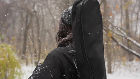 vista lateral de una dama con gorra negra y sudadera con capucha, salpicada de nieve, ajustando la correa de su paquete de guitarra mientras camina a través de un parque cubierto de nieve con árboles cargados de helada