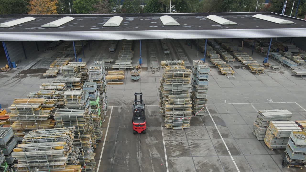 Aerial of a forklift truck driving over an industrial storage yard. The camera tilts up to a large warehouse and reveals high stacks of construction materials