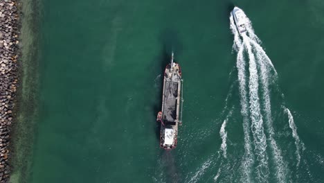 a luxury boat passing a large industrial sand dredging ship next to a coastal seaway rock wall