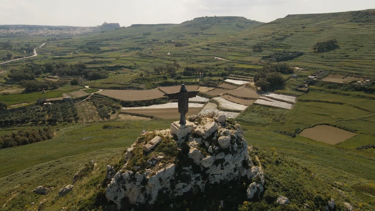estatua de cristo resucitado en gozo, malta, mostrada en un dron circular de 180 grados con el campo verde al fondo