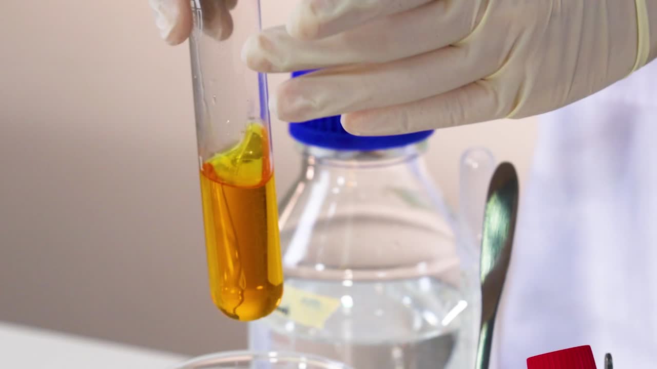 A scientist wearing gloves mixes a yellow solution in a test tube over a beaker.