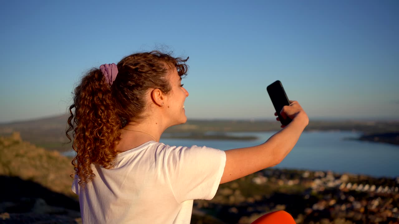 mujer tomando una foto sentada en la ladera de una montaña rocosa bajo un cielo azul