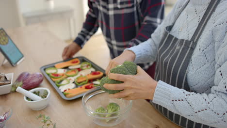 Happy diverse senior couple preparing vegetables and using tablet in kitchen, slow motion