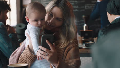 Madre-Joven-Con-Bebé-En-La-Cafetería-Usando-Un-Teléfono-Inteligente-Tomando-Café-Relajándose-En-Un-Restaurante-Concurrido-Disfrutando-De-La-Maternidad