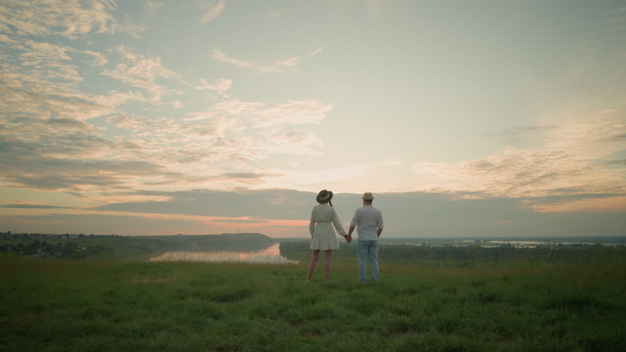 새로 결혼한 부부는 해가 지는 초원 언덕에서 손을 잡고 서서 하늘을 쳐다보고 있다.  셔츠, 모자, 청바지를 입은 남자는  드레스와 모자를 입은 여자와 손을 잡는다.