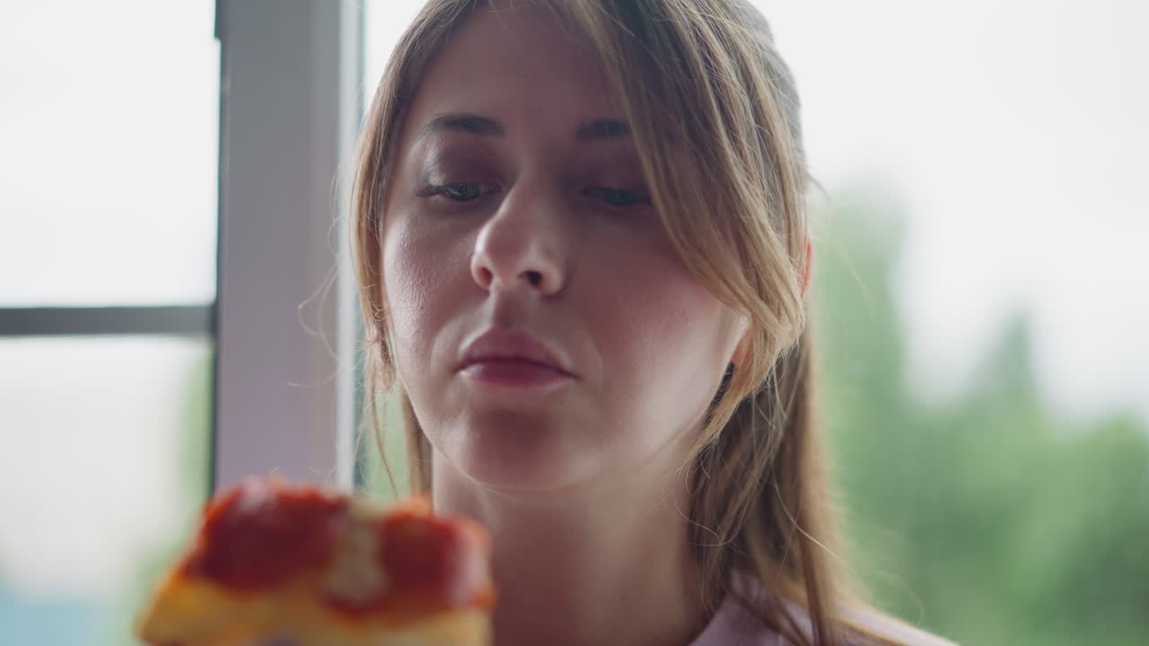Beautiful girl on holiday enjoying grilled egg pizza seated close to window while observing slice with calm expression, natural light illuminating face, greenery blurred outsid