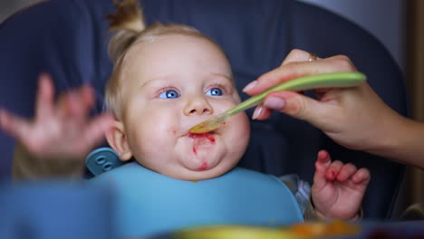 lovely blue-eyed baby with a bib is fed from spoon. close up. nourishing an infant at home.