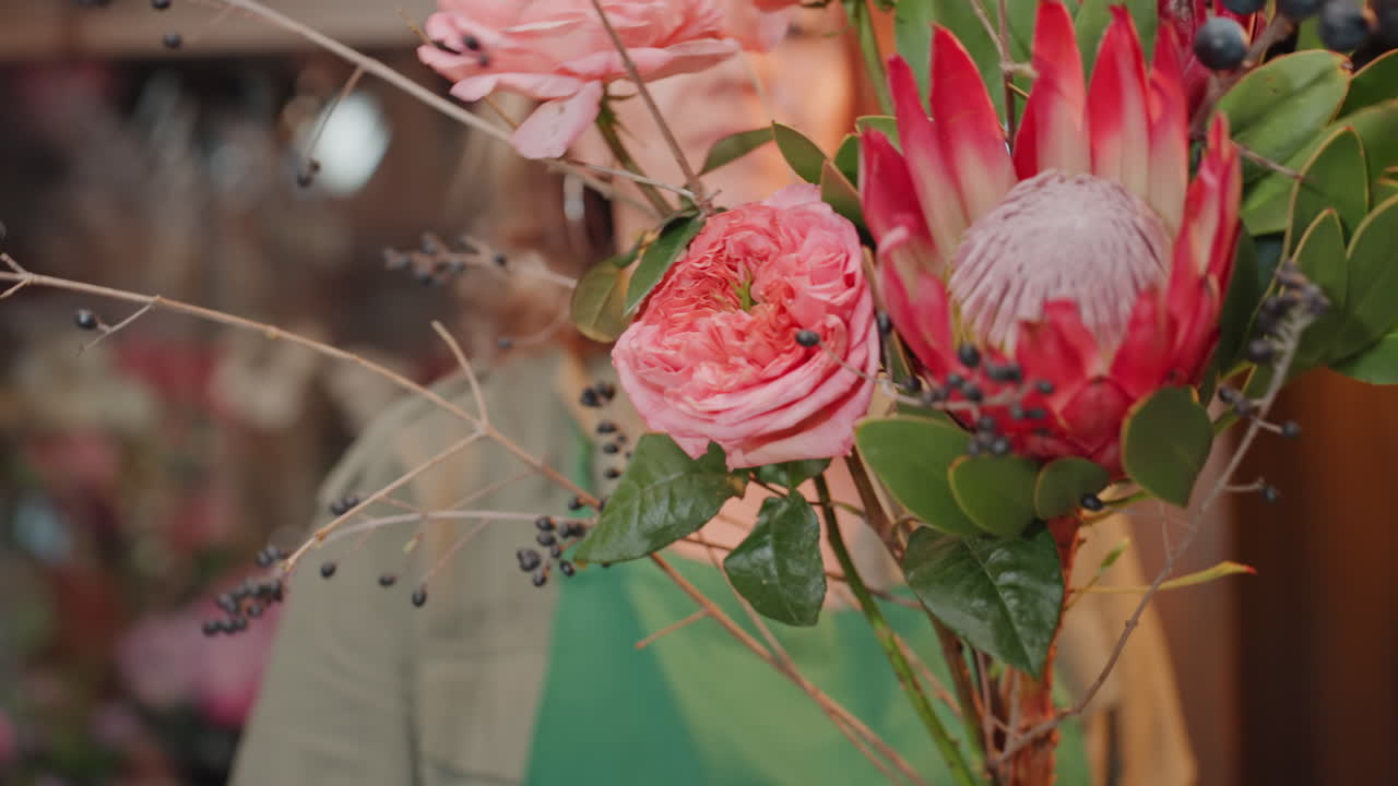 Fiorista bianca ispeziona da vicino un bouquet di protee rosa, con espressione contemplativa mentre verifica la salute degli steli e la consistenza dei petali in un accogliente negozio di fiori, tra luci calde, scaffali rustici e rotoli di nastro, le mani in vista.