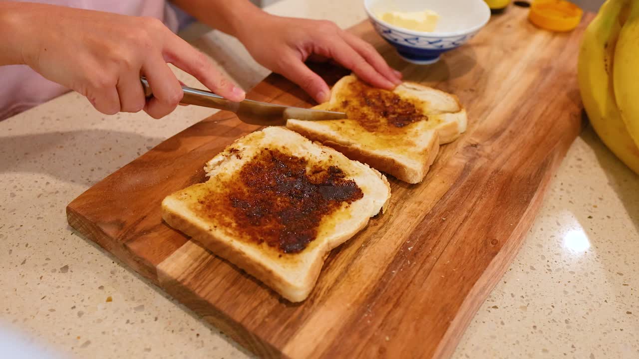 Hands spreading Vegemite on buttered toast slices on a wooden board in a well-lit kitchen
