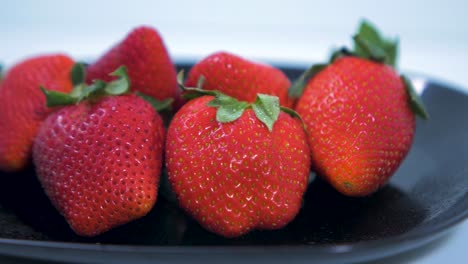 fresh big red tasty ripe strawberries rotates slowly on a black plate on light blue background, healthy food concept, medium close up shot, camera rotate right