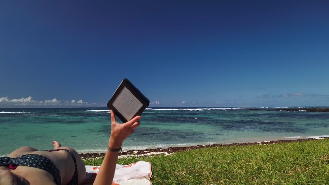 una toma que muestra al máximo a una mujer leyendo un libro electrónico en la playa, con un paisaje asombroso