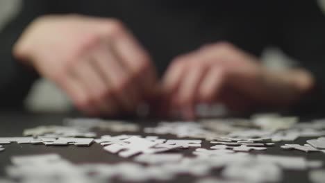 Puzzleteile,-Die-Auf-Einen-Dunklen-Tisch-Fallen-Und-Von-Den-Händen-Einer-Person-Abprallen,-Die-Versucht,-Das-Puzzle-Im-Hintergrund-Zu-Lösen