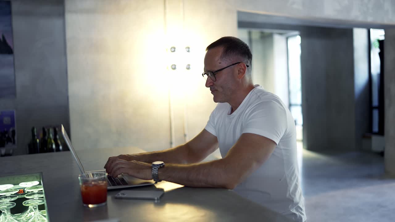 Smiling businessman working on laptop computer at bar. Male professional typing on laptop keyboard at bar workplace. Portrait of positive business man looking at laptop screen indoors