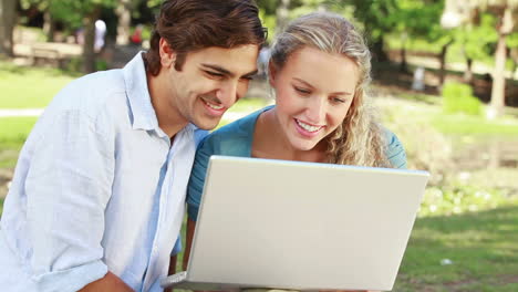 A-happy-couple-in-the-park-with-a-laptop-laughing-as-they-look-to-the-camera