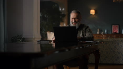 Joyful-old-man-making-video-call-on-computer-in-luxury-cabinet