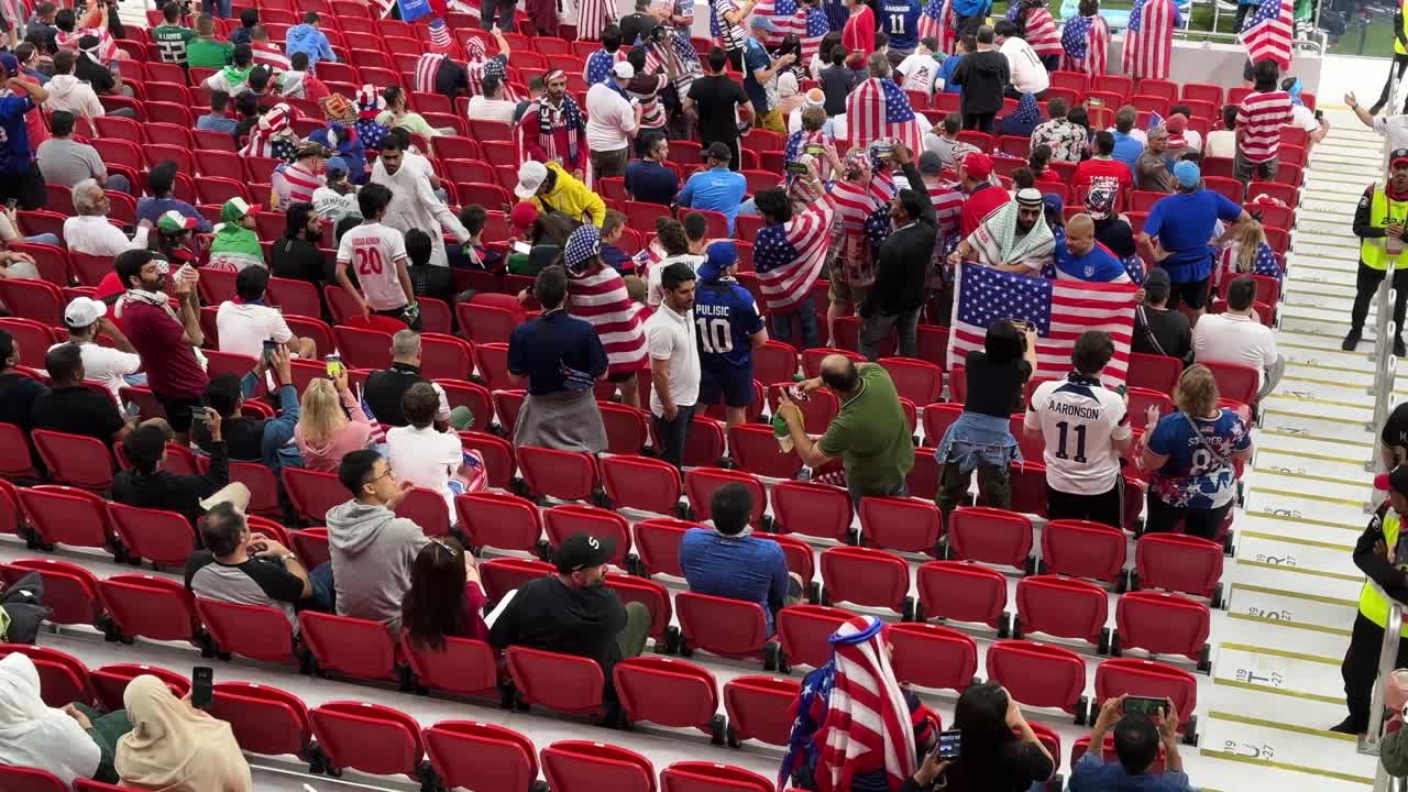 USA Fans in the stadium al thumama are support their football player team with the USA flag in the red seats near the pitch green field of the stadium with beautiful architectural design with friends