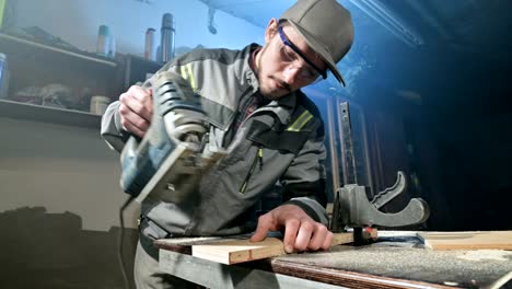 young man with a beard in gray overalls and goggles by profession a carpenter works as an electric jigsaw in his home workshop