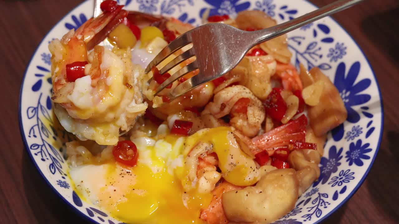 A fork lifts shrimp from a vibrant Thai rice meal with garlic, chili, and runny egg, under warm indoor lighting on a wooden table