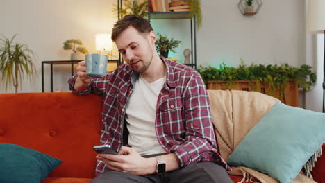Happy-young-man-uses-smartphone-having-chat-on-social-media-while-having-hot-coffee-from-cup-at-home