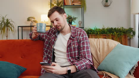 happy young man uses smartphone having chat on social media while having hot coffee from cup at home