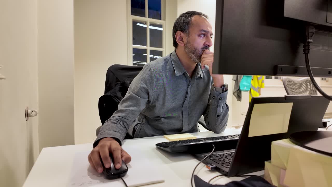 Uomo che lavora al computer in ufficio fino a tardi usando il mouse, guarda lo schermo, indossa una maglietta grigia, con scrivania per laptop e finestra sullo sfondo