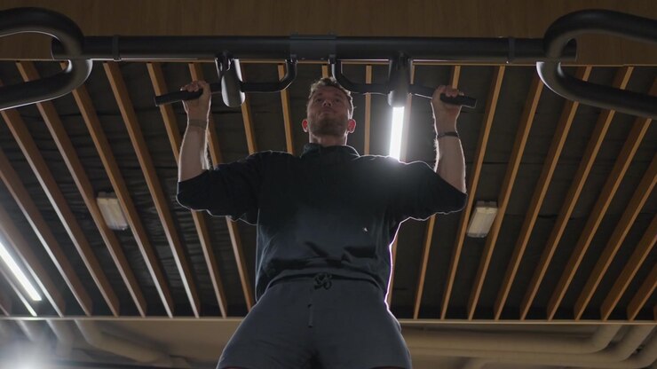 Rear low-angle slow motion view follows an athletic man completing wide grip pull-ups on an overhead bar inside a modern gym.