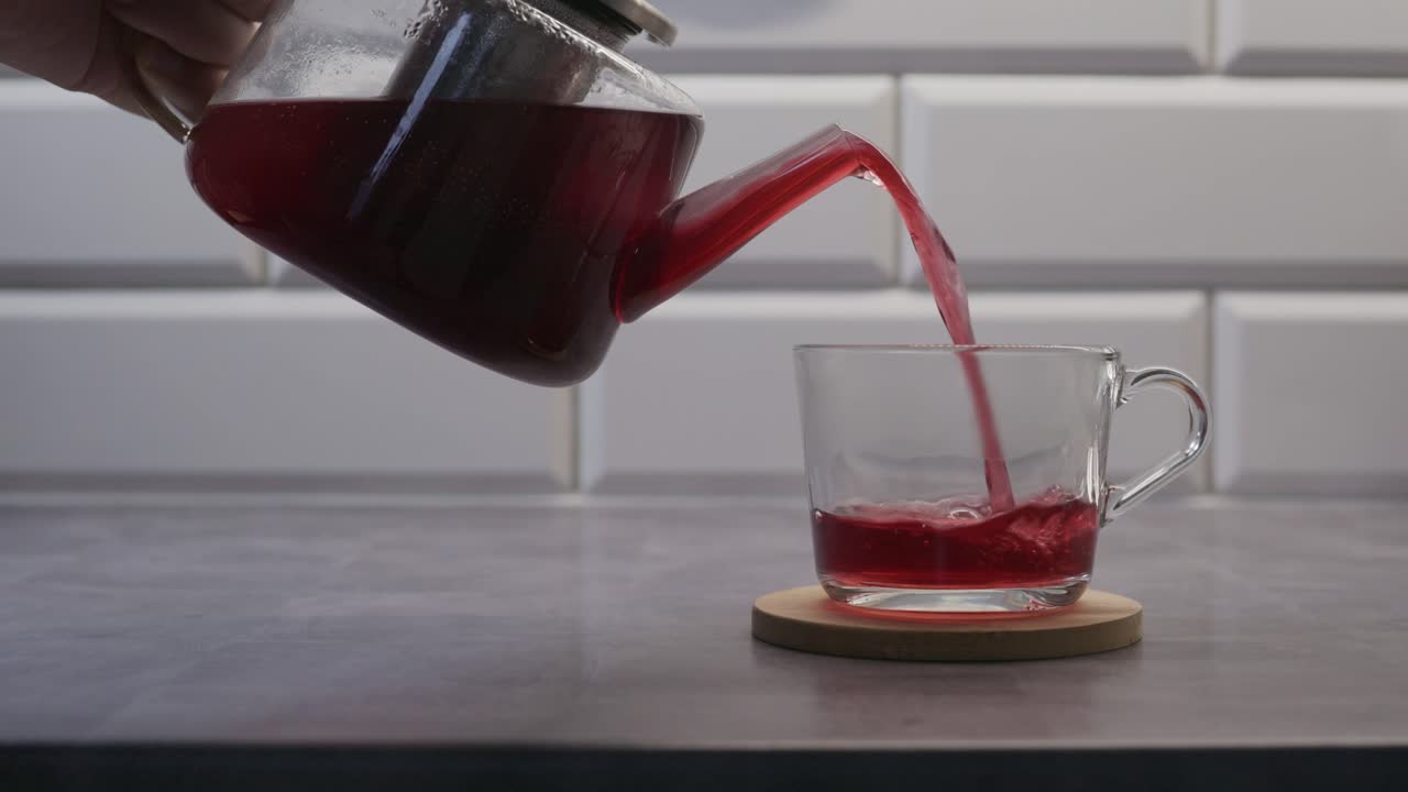 Del tè rosso caldo alle erbe viene versato da una teiera di vetro in una tazza trasparente posizionata su un sottobicchiere di legno. Lo sfondo pulito della cucina e l'angolazione ravvicinata creano una scena rilassante e moderna di preparazione di bevande.