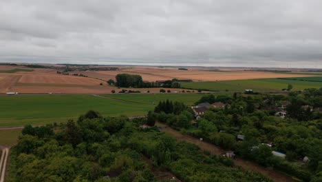 aerial drone view of the french countryside. a highway with traffic cuts through a patchwork of brown and green agricultural fields next to a small village with dense trees under a cloudy sky