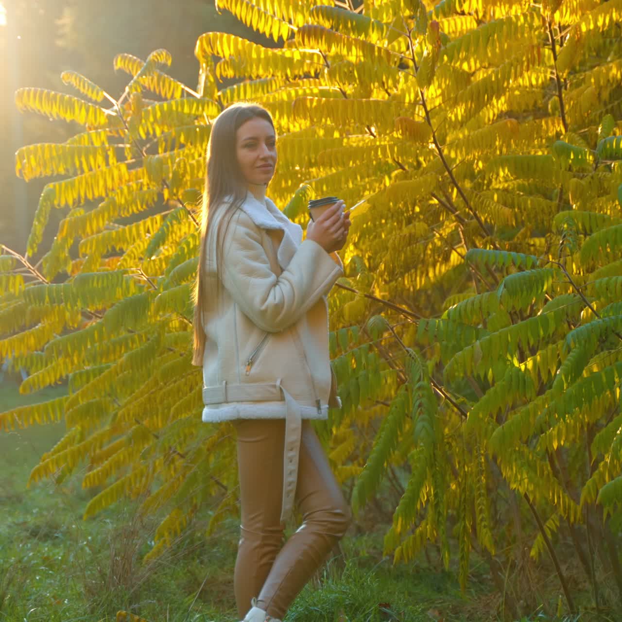 Young woman in white jacket and leggings standing at the big green and yellow bush. Lady is drinking coffee from paper cup then turning to the bright sun showing a maple leaf