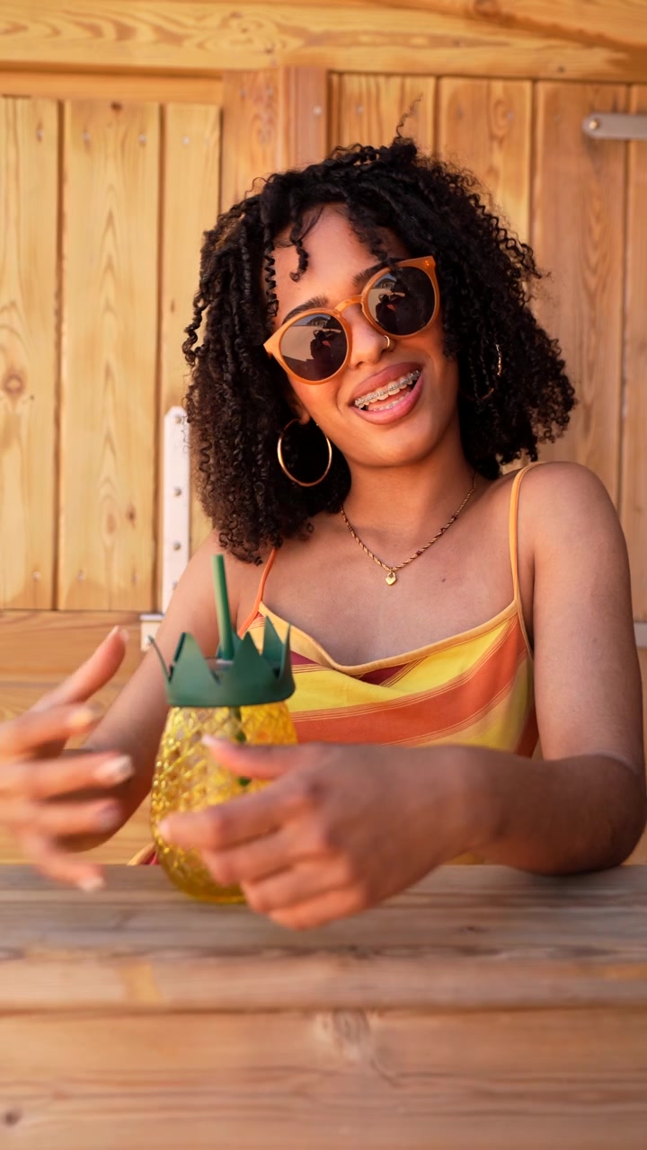 Giovane donna felice con i capelli ricci e gli occhiali da sole che tiene in mano una bevanda all'ananas