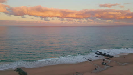 aerial view dusk waves caressing sandy beach under sunset sky. pink clouds hover
