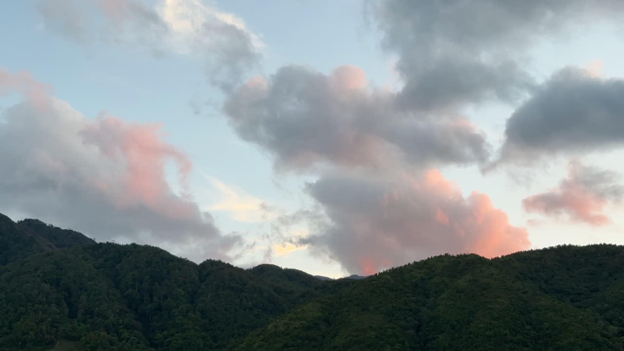 Fixed shot of pink sunset clouds moving quickly above dark mountain peaks. The light contrasts beautifully with the evening landscape.