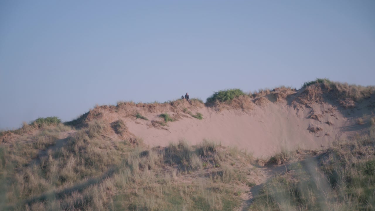 Daytime view of a sandy dune slope covered in tufts of grass, set against a clear blue sky.