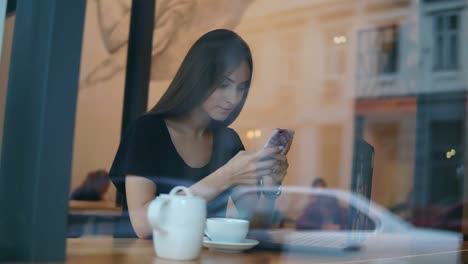 Vista-Desde-El-Exterior-De-Una-Joven-Concentrada-Con-Cabello-Largo-Y-Maquillaje-Natural-Usando-Su-Teléfono-Móvil-En-La-Toma-De-Café