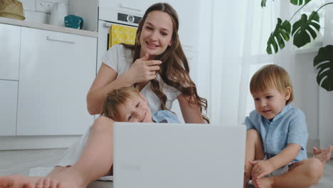 mamá con dos hijos pequeños sentada en el suelo de un apartamento moderno en un interior luminoso con un portátil. mira la pantalla del portátil y mira fotos familiares. haz compras en línea para niños.