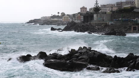 Raue-Meereswellen-Brechen-Gegen-Die-Felsige-Küste-Des-Strandes-Mit-Architekturen-Am-Wasser-Im-Hintergrund-In-Genua,-Italien