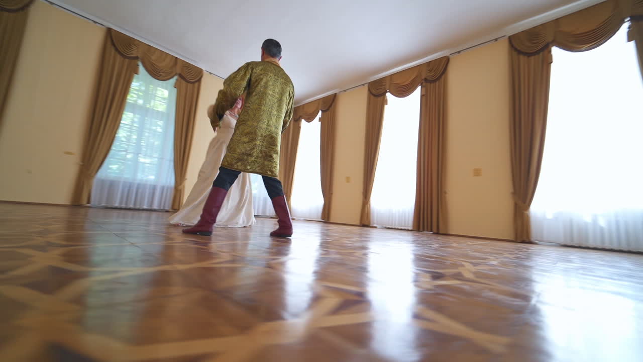 Aged couple wearing old times costumes dancing in the large spacious room. Adult people dancing in the historic atmosphere of a castle. Low angle view.