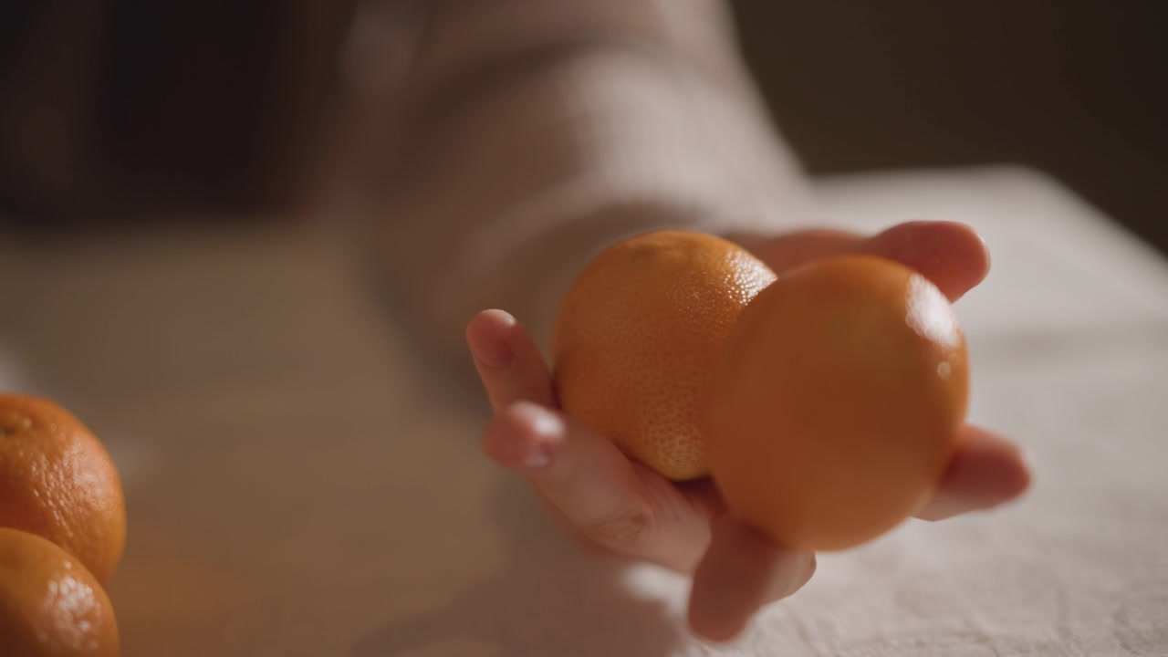 Mão equilibrando laranja na mesa, nutricionista testando a maturação e textura, luz fílmica quente, superfície de linho com tangerinas espalhadas, pontas dos dedos delicadas ajustando a fruta, atmosfera acolhedora.