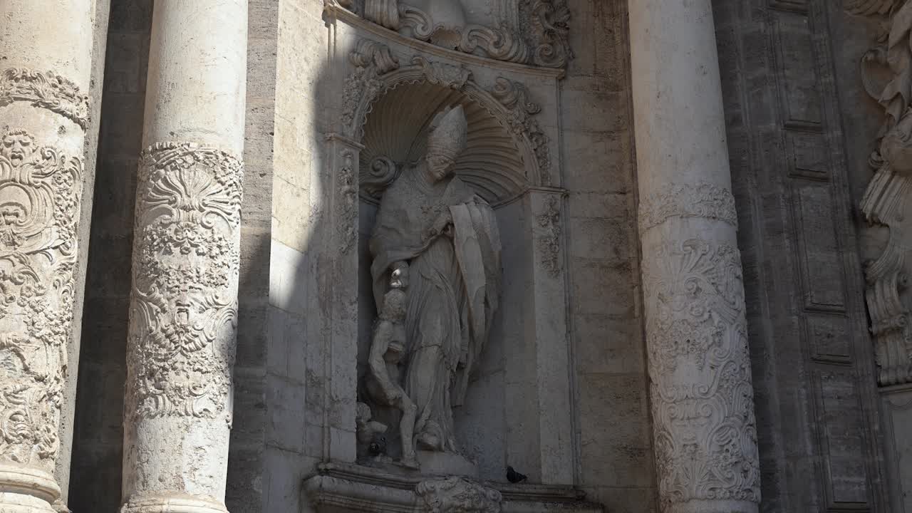 Un monumento in bronzo del Pontefice si erge su un piedistallo fuori dalle storiche mura in pietra della cattedrale gotica sotto un cielo limpido e luminoso.