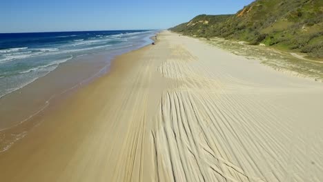 View-along-the-broad-golden-sands-of-Fraser-Island,-with-sharp-shadows-cast-by-the-4x4-tracks-and-a-convoy-of-four-wheel-drive-cars-driving-along-the-beach