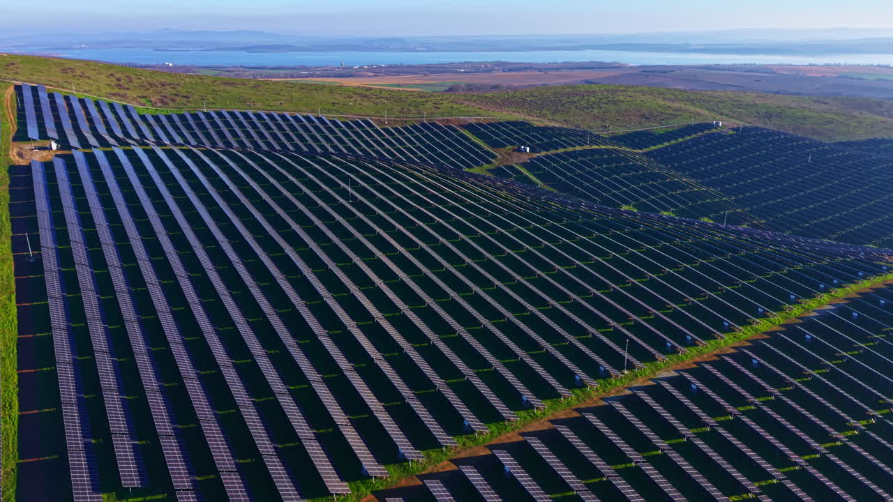Solpaneler dækker bølgende bakker under en klar blå himmel nær et roligt vandområde, hvilket viser fremskridt inden for vedvarende energi i et frodigt grønt landskab.