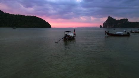 Traditional-Thai-longtail-boats-floating-on-calm-water-at-sunset-with-dramatic-orange-pink-sky-in-the-background-overflight-flyover-drone