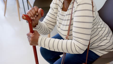 midsection of african american senior woman with cane sitting in hospital waiting room, slow motion